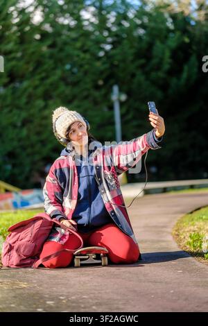 Frau kniet auf einem Skateboard und macht mit einem Smartphone ein Selfie im Park Stockfoto