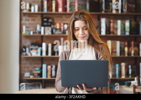 Lächelnde junge Frau arbeitet an einem Laptop in einem gemütlichen Wohnzimmer, modernes Interieur Stockfoto