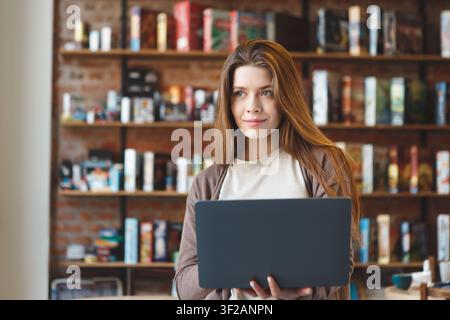 Junge kaukasische Frau, die in einem gemütlichen Buchladen einen Laptop benutzt und sich auf die Arbeit konzentriert Stockfoto