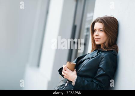 Junge Frau hält Kaffeetasse, lehnt sich an Wand, entspannte Emotion, Lebensstilkonzept Stockfoto