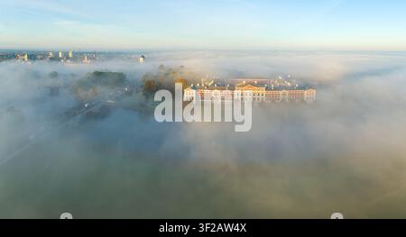 Luftbild des Schlosses Jelgava, umgeben von dichtem Morgennebel, mit einer Brücke über die Lielupe und der Stadt Silhouette in der Ferne in Jelgava, L Stockfoto