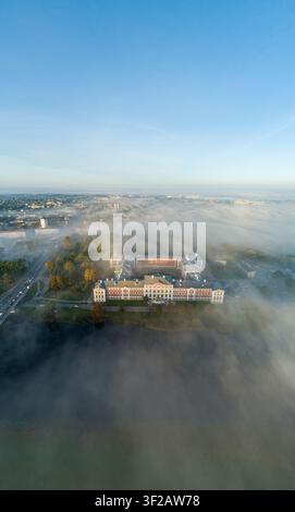 Luftbild des Jelgava-Schlosses, umgeben von dichtem Morgennebel, mit der Lielupe im Vordergrund und klarem blauem Himmel darüber in Jelgava, Lettland. Stockfoto