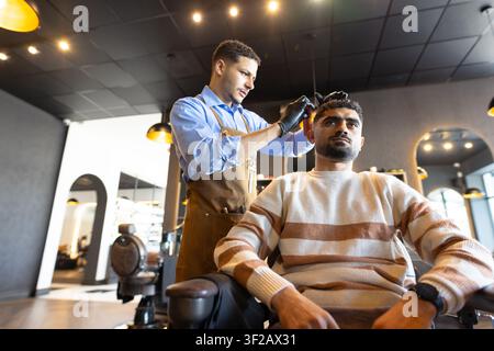 Männlicher Friseur steht hinter männlichem Kunden im Friseurstuhl und schneidet mit einem Haarschneider im Friseursalon die Haare. Pflege, Handwerkskunst, Präzision, zeitgenössisch, mir Stockfoto