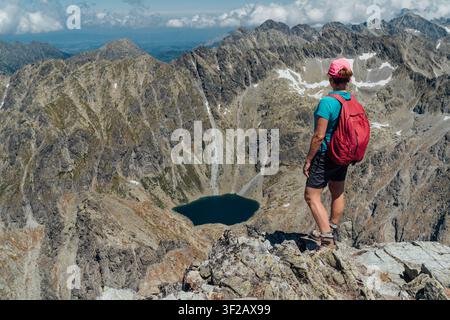 Frauentrekker steht auf dem felsigen Gipfel des Berges Krivan und blickt auf den Bergsee Pleso, weibliche Wanderin mit Rucksack genießt die malerische Aussicht auf die Hohe Tatra Stockfoto