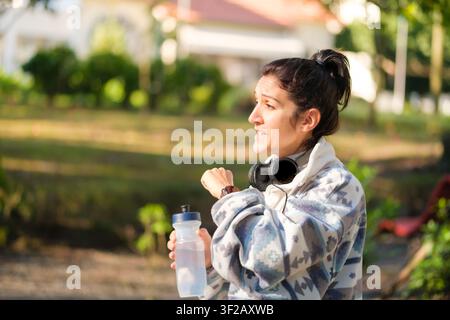 Frau unterbricht Training, überprüft Smartwatch und hält eine Wasserflasche im Park Stockfoto