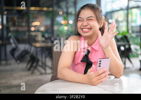 Eine fröhliche junge südostasiatische thailändische Isan-Frau winkt mit der Hand, während sie ihr Smartphone an einem Außentisch eines Cafés hält. Sie hat ein lustiges, fröhliches Gesicht Stockfoto