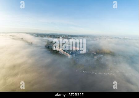 Luftaufnahme des Jelgava-Palastes, umgeben von dichtem Morgennebel, mit der umliegenden Stadt und dem Fluss in der Ferne unter einem klaren blauen Himmel in J Stockfoto