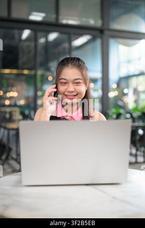 Eine vertikale Mittelaufnahme einer fröhlichen südostasiatischen thailändischen Isan-Frau, die herzlich lächelt, während sie an ihrem Smartphone spricht, hinter ihrem Laptop an einem Cafétisch. Stockfoto