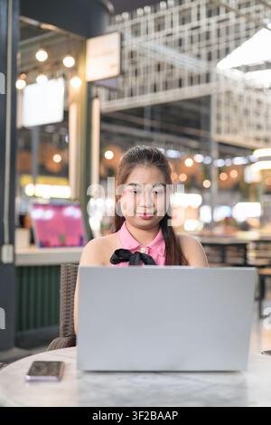 Eine vertikale Mittelaufnahme, die sich auf eine südostasiatische thailändische Isan-Frau konzentriert, die auf ihren silbernen Laptop-Bildschirm schaut, während sie in einem Café sitzt. Der Hintergrund zeigt Stockfoto