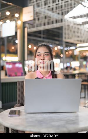 Eine vertikale Mittelaufnahme einer entspannten südostasiatischen thailändischen Isan-Frau, die mit einem friedlichen Ausdruck aus der Kamera schaut, während sie hinter ihrem Laptop sitzt Stockfoto