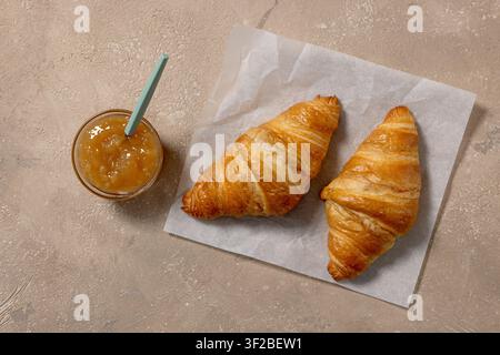 Oberansicht von zwei frisch gebackenen Buttercroissants, serviert mit einer Schale Fruchtschmier auf einem beigen Küchentisch. Gemütliche Frühstückskomposition mit blättrigem Stockfoto