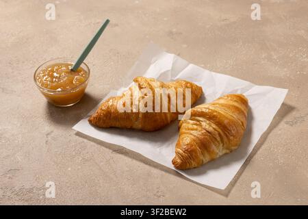 zwei frisch gebackene Buttercroissants, serviert mit einer Schüssel Fruchtmarmelade auf einem beigen Küchentisch. Gemütliche Frühstückskomposition mit goldener, blättriger Pastete Stockfoto