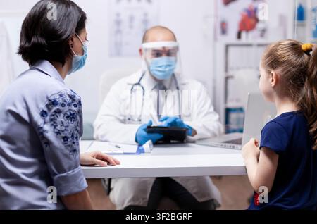 Kinderarzt mit Gesichtsmaske nutzt ein Tablet während der Beratung eines Kindes und zeigt pädiatrische Gesundheitsversorgung und digitale medizinische Unterstützung. Stockfoto