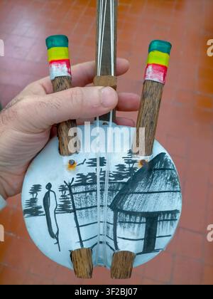Eine Nahaufnahme von vorne eines handgefertigten afrikanischen Saiteninstruments, das von einer Hand gehalten wird. Der runde Resonanzkörper zeigt eine schwarz-weiß handgemalte Szene Stockfoto
