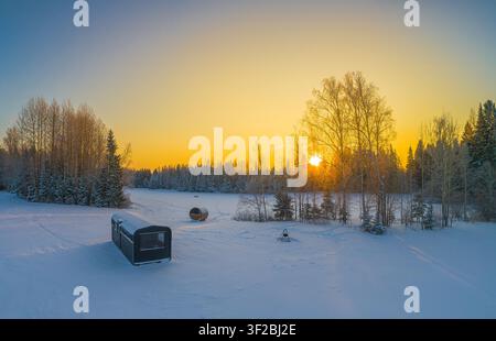 Luftaufnahme einer kleinen Hütte und einer Fasssauna in einem schneebedeckten Feld, umgeben von einem Wald während eines goldenen Sonnenuntergangs in Lettland. Stockfoto