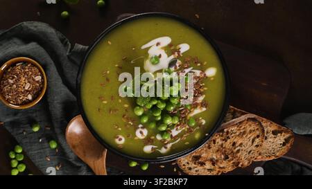Eine Top-Down-Studioaufnahme zeigt eine keramische Schüssel gefüllt mit lebendiger grüner Erbsensuppe, garniert mit Sahne und Leinsamen. Stimmungsvolle Beleuchtung betont die Texturen Stockfoto