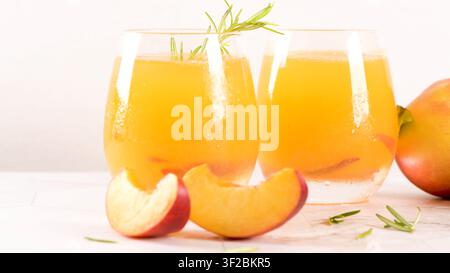 Eine Augenhöhe-Aufnahme zeigt zwei stiellose Gläser, gefüllt mit lebhaftem gelbem Pfirsichsaft, dekoriert mit einem Zweig frischem Rosmarin. Scheiben von saftigen Pfirs Stockfoto