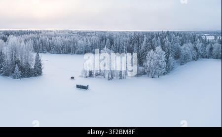Luftaufnahme einer kleinen dunklen Hütte in einem weiten schneebedeckten Feld, umgeben von frostbedeckten Bäumen und einem dichten Wald in Lettland. Stockfoto