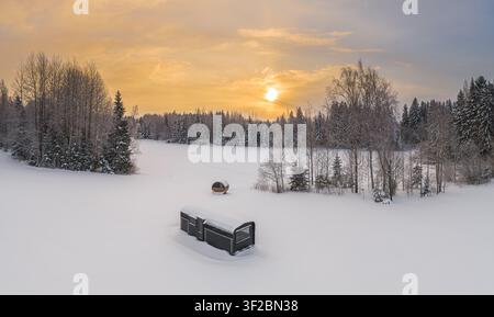 Luftaufnahme einer kleinen Hütte und einer Fasssauna in einem schneebedeckten Feld, umgeben von einem Winterwald bei Sonnenuntergang in Lettland. Stockfoto