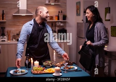 Ehefrau lächelt ihren Ehemann im Esszimmer an, während er sie mit einem romantischen Abendessen, gesunden Speisen und einem festlichen Tisch überrascht, wobei der unterstützende Feiertags-, Ess- oder Beziehungszusammenhang sichtbar bleibt. Stockfoto