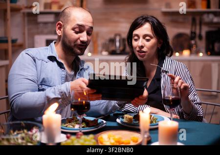 Fröhliches junges Paar isst zusammen und nutzt dabei ein Tablet während eines romantischen Abendessens, kombiniert Internetsurfen mit dem Jahrestag. Stockfoto
