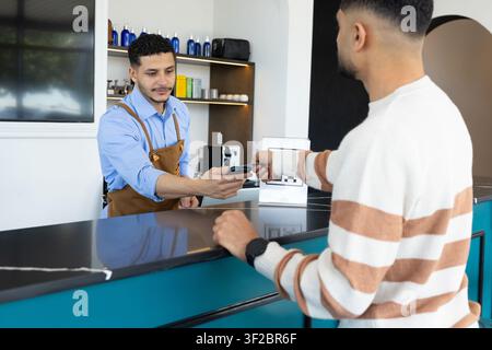 Afrikanischer männlicher Kunde, der Karte antippt, männlicher Barista in Schürze, der Terminal an türkisfarbenem Tresen anbietet Stockfoto