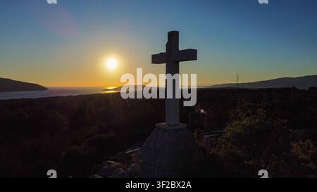 Weißes Steinkreuz-Denkmal auf dem Hügel der Insel Hvar, das einen panoramischen Sonnenuntergang über die Bucht von Stari Grad bietet, Kroatien Stockfoto