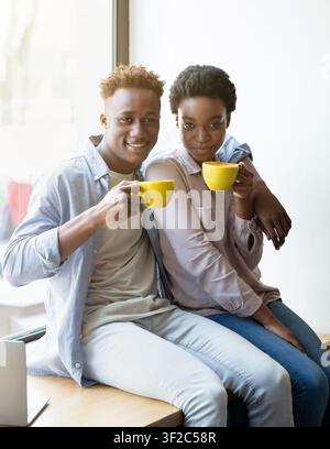 Positives schwarzes Paar sitzt auf Fensterbank und trinkt aromatischen Kaffee Im Café Stockfoto