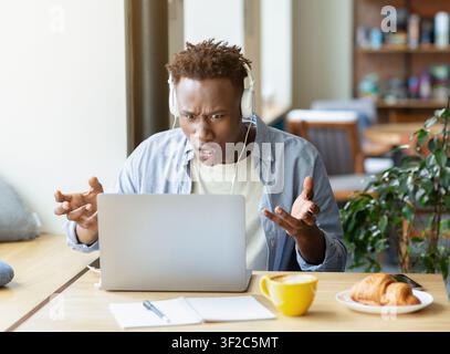 Gestresster Afroamerikaner in Kopfhörern, der Laptop benutzt, um an komplizierten Geschäftsprojekten zu arbeiten, und mit Problemen im städtischen Café konfrontiert. Junger schwarzer Student ha Stockfoto