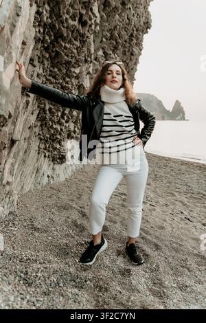 Frau Strandmode posiert in Lederjacke und gestreiftem Pullover mit Kopierraum bei Felsformation an der Küste Stockfoto