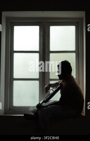 Eine Silhouette eines Musikers, der am Fenster sitzt und Gitarre spielt. Die ruhige Szene vermittelt ein Gefühl von Gelassenheit und Selbstreflexion, verstärkt durch so Stockfoto