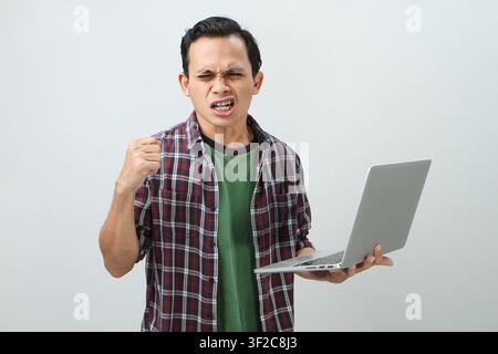 Wütender verrückter asiatischer indonesischer Mann, der einen Laptop-Computer auf isoliertem Hintergrund hält Stockfoto