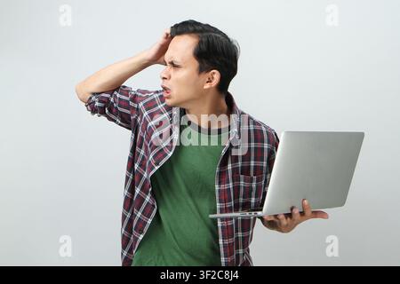 Wütender verrückter asiatischer indonesischer Mann, der einen Laptop-Computer auf isoliertem Hintergrund hält Stockfoto