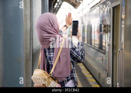 Rückansicht einer asiatischen muslimischen Frau, die ein Smartphone hält und auf dem Bahnhofsgleis zum Abschied winkt. Reisekonzept Stockfoto