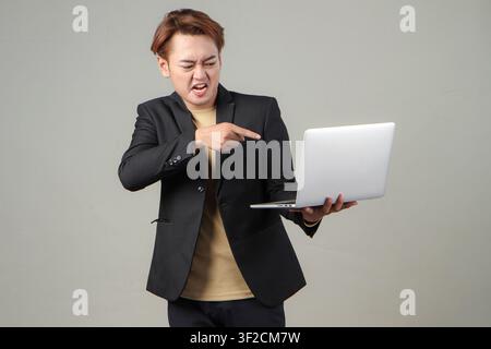 Porträt eines wütenden asiatischen Geschäftsmannes, der einen Laptop hält, einen Anzug trägt, vor isolierendem Hintergrund Stockfoto