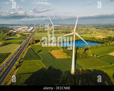 Drohnenaufnahme einer Windturbine, die einen langen Schatten über grüne Felder neben einem blauen Teich und einer Autobahn wirft, eine Sommerm Szene nachhaltiger Windenergie Stockfoto
