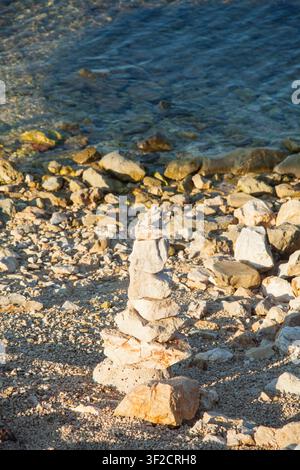 Ein ausgewogener Stapel aus Natursteinen an einem felsigen Strandufer mit klarem blauem Wasser im Hintergrund, der Ruhe, Balance und Natur symbolisiert. Stockfoto