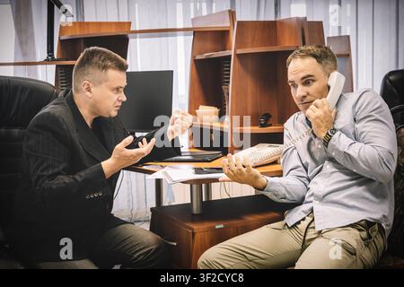 Zwei empörte Männer streiten am Telefon im Büro, versuchen, ihren Standpunkt zu beweisen, intensive Verhandlung. Stockfoto
