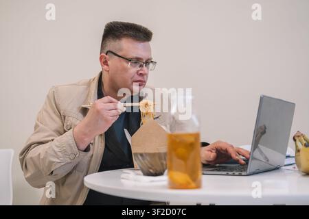 Konzentrierter Geschäftsmann isst mit Stäbchen Take-out-Nudeln, während er an einem Laptop arbeitet, und verbindet Mittagspause und Fernarbeit an einem Home-Office-Schreibtisch. Stockfoto