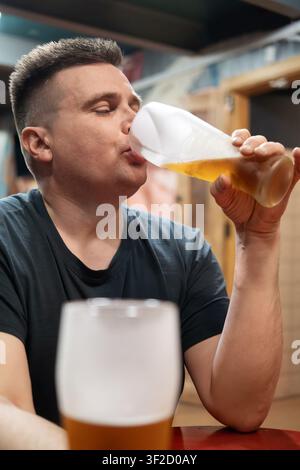 Müder Mann in einem dunklen T-Shirt, der Bier aus einem großen Glasmug mit weißem Schaum trinkt, sitzt an einem Bartisch. Stockfoto