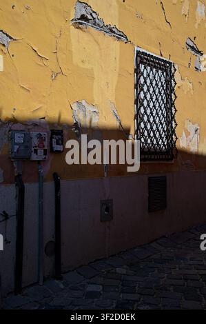 Szentendre, Ungarn - 04. Juli 2026: Gelbe verwitterte Wand mit abblätternder Farbe, vergittertes Fenster mit Eisengitter und Schatten auf dem Kopfsteinpflaster. Stockfoto