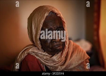eine ältere Frau, die einen hellfarbenen Kopftuch trägt, sitzt im Inneren in weichem natürlichem Licht. Stockfoto