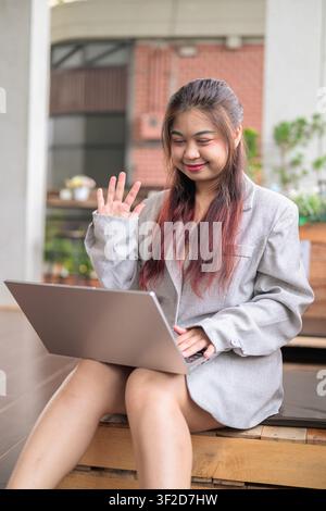 Eine vertikale Mittelaufnahme einer jungen südostasiatischen thailändischen Isan-Geschäftsfrau, die einen offenen Laptop hält und warm lächelt, während sie mit der Hand zum Bildschirm winkt Stockfoto