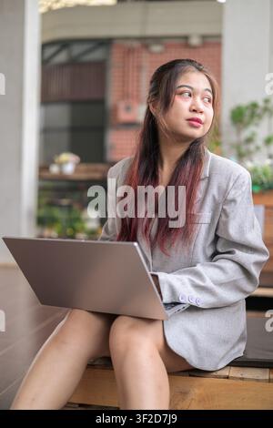 Eine vertikale Mittelaufnahme einer jungen südostasiatischen thailändischen Isan-Geschäftsfrau, die im Freien sitzt, mit einem silbernen Laptop aufgeschlagen auf ihrem Schoß, und die weg von ihr schaut Stockfoto