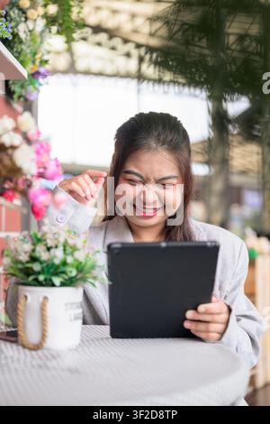 Eine vertikale Mittelaufnahme einer jungen südostasiatischen thailändischen Isan-Geschäftsfrau, die an einem Café-Tisch sitzt, die Augen schließt und mit einer amüsierten, fröhlichen Geste lacht Stockfoto