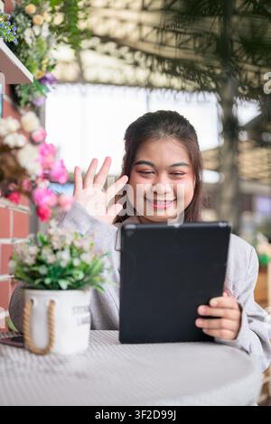 Eine vertikale Mittelaufnahme einer jungen thailändischen Isan-Geschäftsfrau südostasiatischer Herkunft, die im Freien sitzt, warm lächelt und mit der Hand auf ihren Tablet-Bildschirm deutet Stockfoto