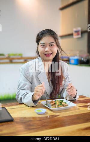 Eine vertikale Mittelaufnahme einer jungen südostasiatischen thailändischen Isan-Geschäftsfrau, die ihr Besteck hält und warm in die Kamera lächelt, bereit, einen Teller zu genießen Stockfoto