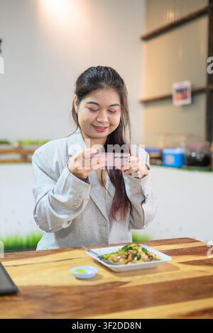 Eine vertikale Mittelaufnahme einer jungen thailändischen Geschäftsfrau aus Isan, die sanft lächelt, während sie ihr rosa Smartphone horizontal hält, um ein Bild zu machen Stockfoto