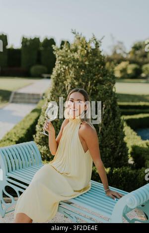 Eine Frau in einem hübschen gelben Kleid sitzt auf einer blauen Bank, lächelt und genießt ein Getränk in einem sonnigen Garten voller Grünpflanzen. Stockfoto