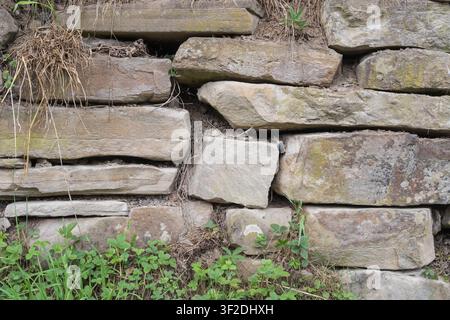 Traditionelle Stützmauer aus Natursteinblöcken mit Wildpflanzen und Gras. Rustikales Mauerwerk und Detail der Landschaft. Stockfoto
